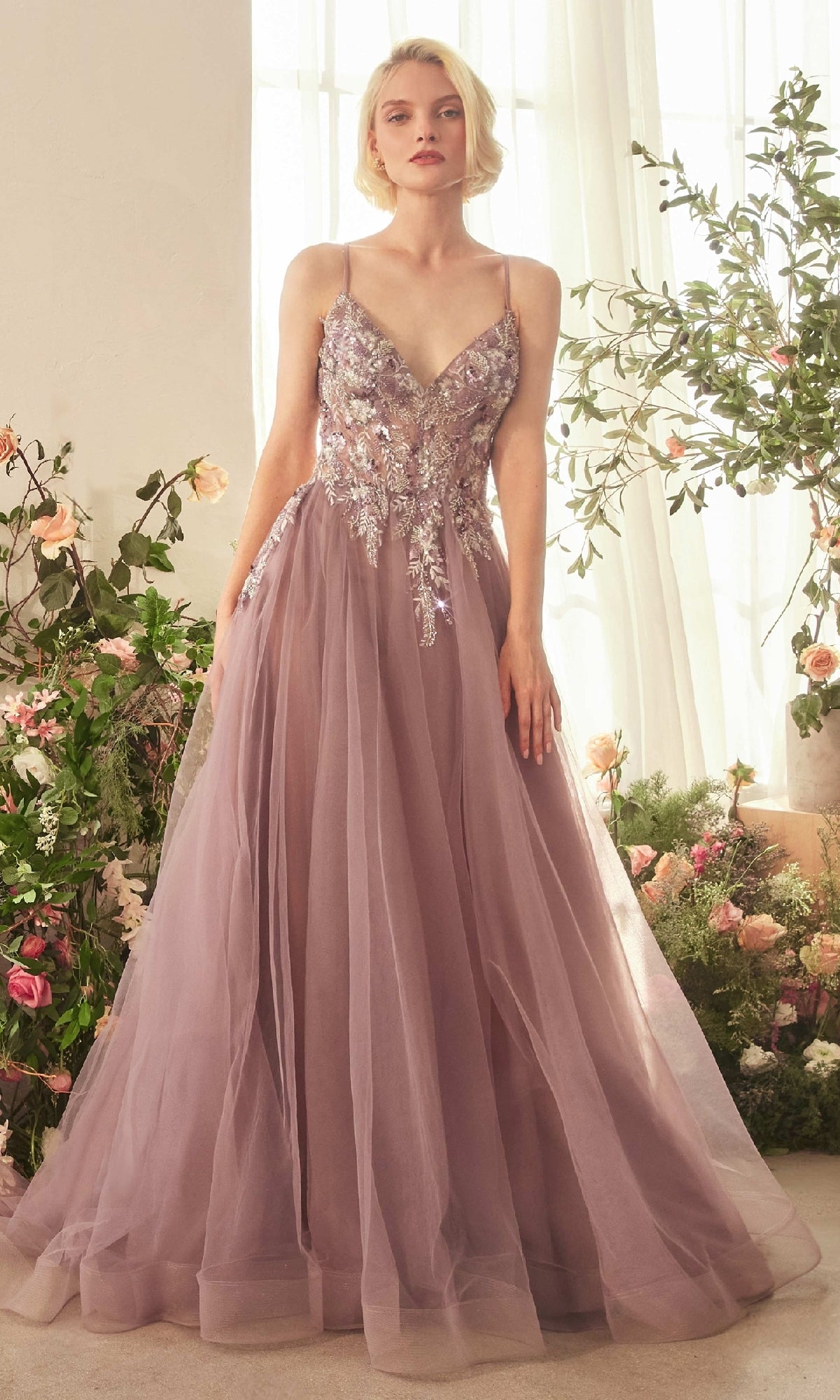 A woman stands indoors near flowers and large windows, wearing the Andrea & Leo A1419 Violet Purple Long Prom Dress with a sequin lace bodice and floral embroidery.